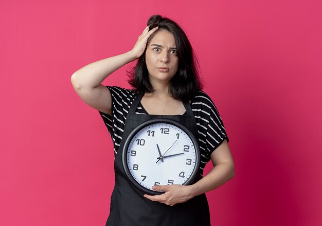 Giovane bella donna parrucchiere in grembiule tenendo l'orologio da parete con la mano sulla sua testa per errore cercando confuso standinh su rosa