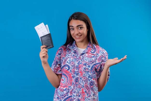 Giovane bella donna piacevole del viaggiatore con i biglietti e passaporto che sembra presentazione sorridente sicura con il braccio della mano che controlla parete blu