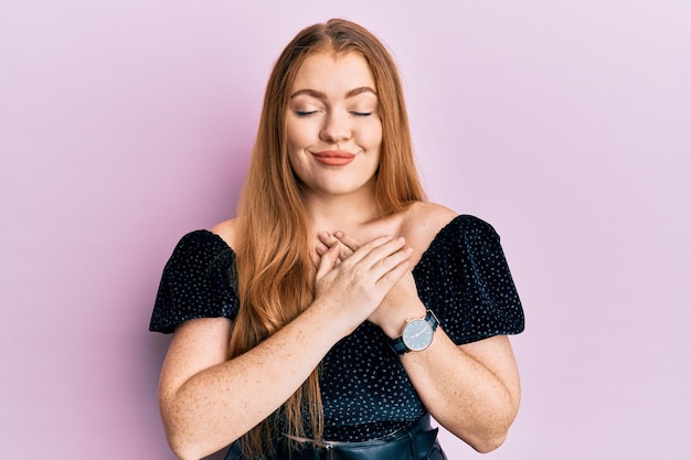 Giovane bella donna rossa che indossa un look elegante e sexy sorridente con le mani sul petto, gli occhi chiusi con gesto grato sul viso. concetto di salute.