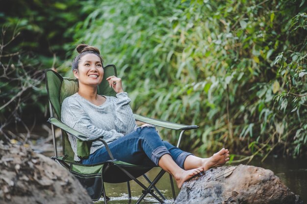 Giovane bella donna seduta su una sedia da campeggio in ruscello per il relax sorride nella foresta naturale durante il viaggio in campeggio con lo spazio della copia di felicità