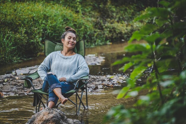 Giovane bella donna seduta su una sedia da campeggio nel ruscello e chiude gli occhi per rilassarsi sorride nella foresta naturale durante il viaggio in campeggio con lo spazio della copia di felicità