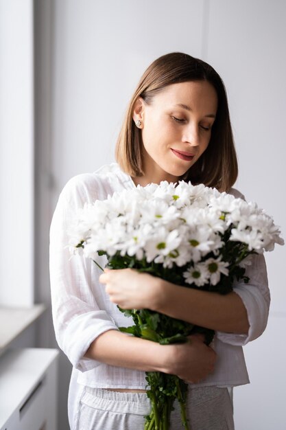Giovane bella donna sorridente adorabile dolce sveglia con tenere un mazzo di fiori freschi bianchi a casa