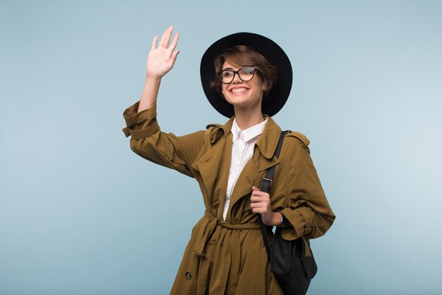 Giovane bella donna sorridente con capelli corti scuri in trench coatocchiali da vista e cappello con zaino che guarda felicemente da parte e sventola su sfondo blu isolato