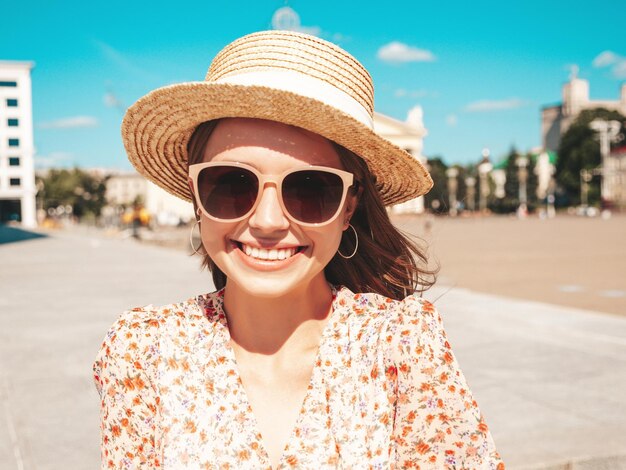 Giovane bella donna sorridente hipster in abiti estivi alla moda Donna spensierata sexy in posa sullo sfondo della strada in cappello da spiaggia al tramonto Modello positivo all'aperto Allegro e felice in occhiali da sole