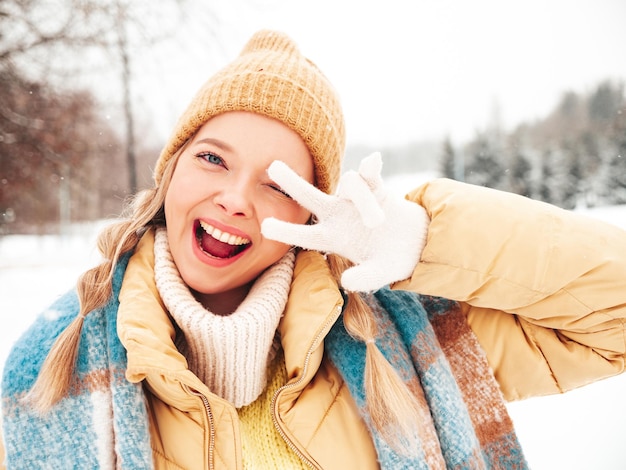 Giovane bella donna sorridente hipster in vestiti caldi alla moda e sciarpa. Donna spensierata in posa in strada nel parco. Modello puro positivo divertendosi nella neve. Godersi i momenti invernali. Mostra il segno della pace