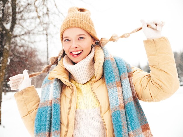 Giovane bella donna sorridente hipster in vestiti caldi alla moda e sciarpa. Donna spensierata in posa in strada nel parco. Modello puro positivo divertendosi nella neve. Godersi i momenti invernali. Mostra la lingua