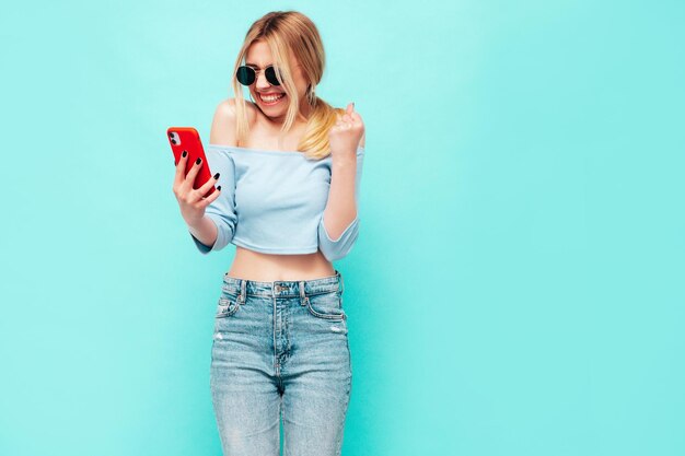 Giovane bella donna sorridente in abiti estivi alla moda Donna eccitata e stupita che posa vicino al muro blu in studio Modello scioccato e fortunato che legge il dispositivo moderno che celebra tenendo il telefono