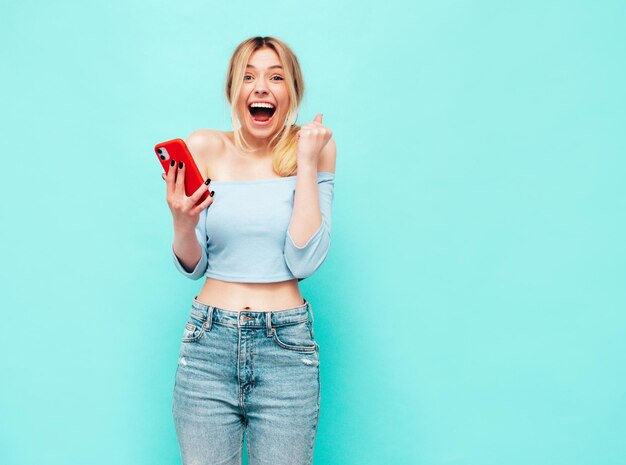 Giovane bella donna sorridente in abiti estivi alla moda Donna eccitata e stupita che posa vicino al muro blu in studio Modello scioccato e fortunato che legge il dispositivo moderno che celebra tenendo il telefono