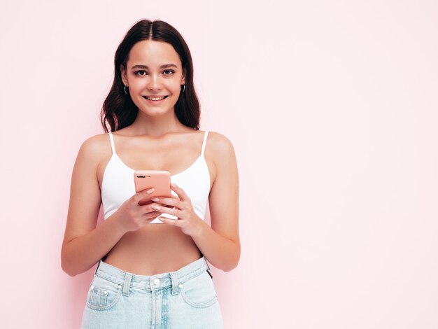 Giovane bella donna sorridente in abiti estivi alla moda Donna spensierata sexy in posa vicino al muro rosa in studio Modello bruna positivo guardando lo schermo del cellulare Holding smartphone e utilizzo di app