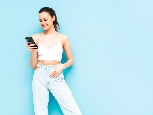 Giovane bella donna sorridente in jeans estivi alla moda Donna spensierata sexy in posa vicino al muro blu in studio Modello bruna positivo guardando lo schermo del cellulare Holding smartphone e utilizzo di app