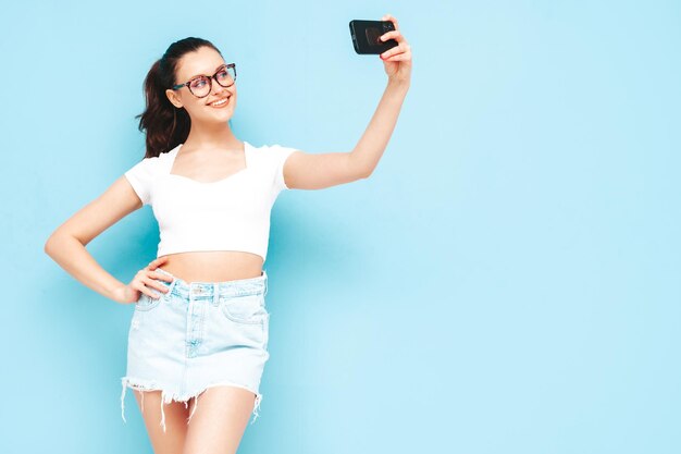 Giovane bella donna sorridente in jeans estivi alla moda gonna e top vestiti Donna spensierata sexy in posa vicino al muro blu in studio Modello bruna positivo divertendosi prendendo selfie