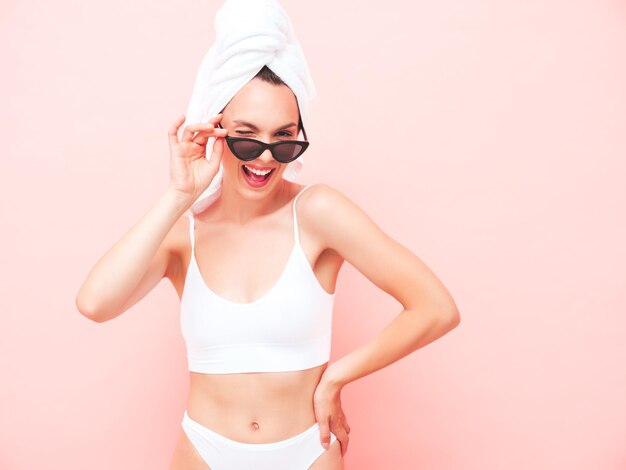 Giovane bella donna sorridente in lingerie bianca Modello spensierato sexy in biancheria intima e asciugamano sulla testa in posa parete rosa in studio Femmina positiva e felice godendo la mattina in occhiali da sole