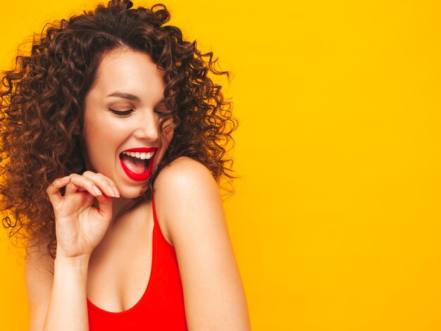 Giovane bella donna sorridente in posa vicino al muro giallo in studioModello sexy in costume da bagno rosso da bagnoFemmina positiva con acconciatura a riccioliFelice e allegra