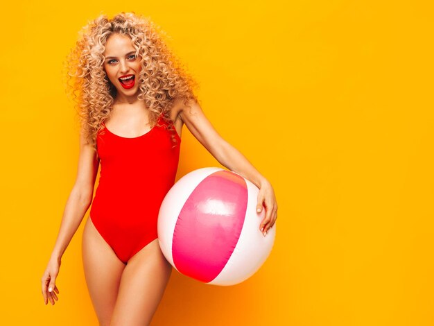 Giovane bella donna sorridente in posa vicino al muro giallo in studioModello sexy in costume da bagno rosso da bagnoFemmina positiva con l'acconciatura di riccioli che tiene palla gonfiabile del pennyFelice e allegro