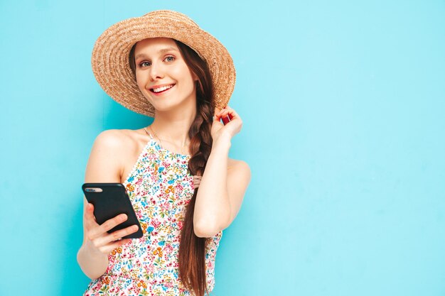 Giovane bella donna sorridente in tuta estiva alla moda Donna sexy con in posa vicino al muro giallo in studio Modello positivo che si diverte con occhiali da sole e cappello Guardando smartphone utilizzando le app