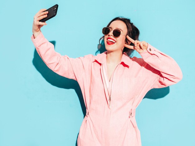 Giovane bella donna sorridente in tuta rosa estiva alla moda Donna spensierata sexy in posa vicino al muro blu in studio con due corna Modello positivo divertendosi e impazzendo Scattare foto selfie