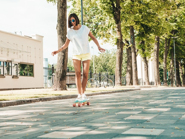 Giovane bella donna sorridente sexy dei pantaloni a vita bassa in occhiali da sole. Ragazza alla moda in maglietta e shorts di estate. Femmina positiva che guida il pattino blu del penny nella via