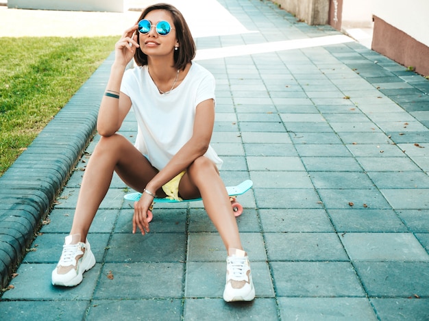 Giovane bella donna sorridente sexy dei pantaloni a vita bassa in occhiali da sole Ragazza alla moda in maglietta e shorts di estate Femmina positiva con il pattino blu del penny che posa sui precedenti della via