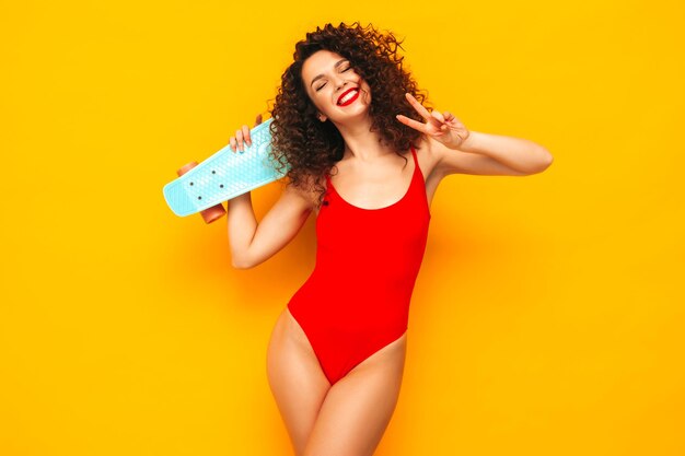 Giovane bella donna sorridente sexy in posa vicino al muro giallo in studioModello alla moda in costume da bagno rosso estivo da bagnoFemmina positiva con riccioli afro Felice e allegro Holding penny skateboard