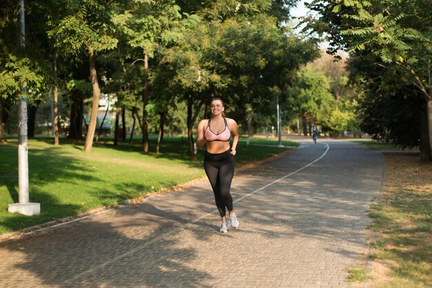 Giovane bella donna taglie forti in top sportivo e leggings che corre felicemente mentre trascorre del tempo nell'accogliente parco cittadino