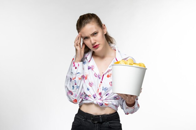 Giovane bella femmina di vista frontale che mangia le patatine fritte e che guarda film ha sottolineato sulla superficie bianca