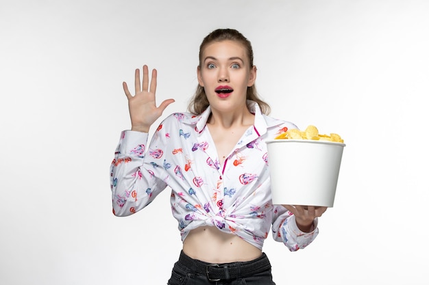 Giovane bella femmina di vista frontale che mangia le patatine fritte e che guarda film sullo scrittorio bianco