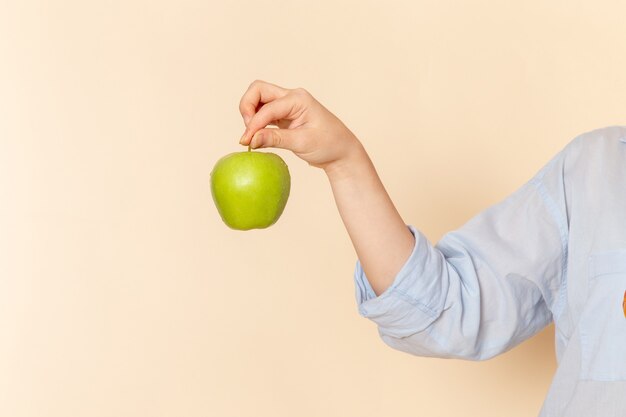 Giovane bella femmina di vista frontale in camicia che tiene mela verde fresca sulla posa della donna del modello della frutta della parete crema