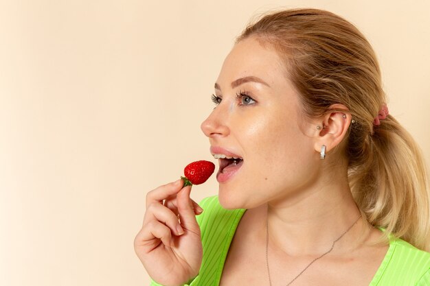 Giovane bella femmina di vista frontale in camicia verde che mangia fragola sulla posa della donna del modello della frutta della parete crema leggera
