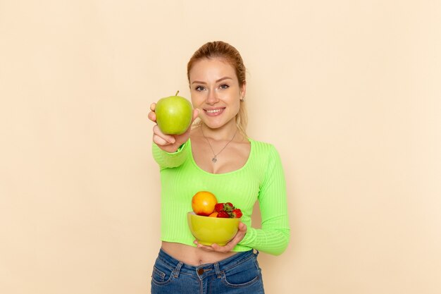 Giovane bella femmina di vista frontale in camicia verde che tiene il piatto con i frutti e che tiene la mela verde sulla posa della donna del modello della frutta della parete crema chiara