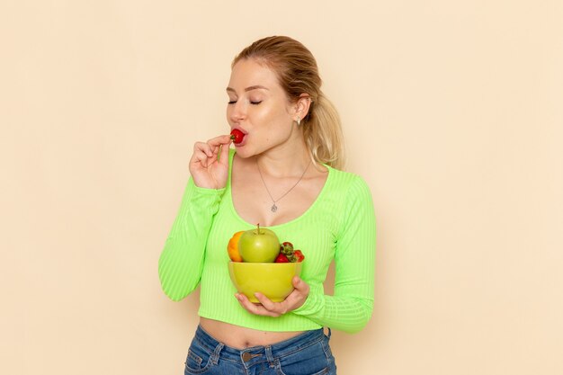 Giovane bella femmina di vista frontale in camicia verde che tiene il piatto con la frutta che mangia fragola sulla posa della donna del modello della frutta della parete crema chiara