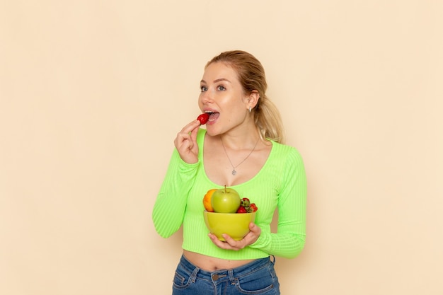 Giovane bella femmina di vista frontale in camicia verde che tiene il piatto con la frutta che mangia fragola sulla posa della donna del modello della frutta della parete crema chiara