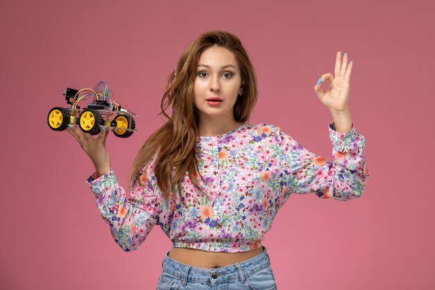 Giovane bella femmina di vista frontale in fiore progettato camicia e blue jeans che tengono automobile del giocattolo in posa sullo sfondo rosa