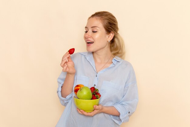 Giovane bella femmina di vista frontale nella zolla della tenuta della camicia con la frutta che mangia sulla posa matura della donna di modello della frutta della parete crema