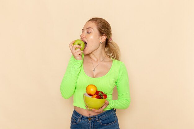 Giovane bella femmina di vista frontale nella zolla verde della tenuta della camicia con i frutti che mangiano mela sulla posa della donna del modello della frutta della parete crema chiara