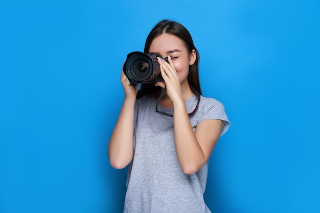 Giovane bella fotografa asiatica focalizzata sulla fotocamera DSLR professionale e sulla parete blu