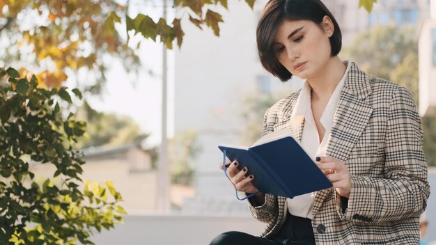 Giovane bella imprenditrice vestita con blazer plaid che redige un business plan per un mese su una strada cittadina Ragazza attraente con blocco note all'aperto