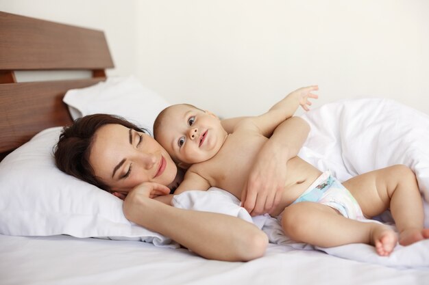 Giovane bella madre con gli occhi chiusi e il suo neonato sdraiato nel letto la mattina presto.