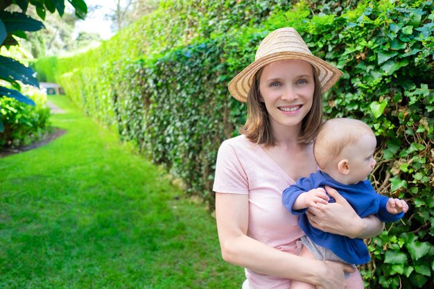 Giovane bella mamma in piedi nel parco, tenendo la figlia, sorridente e guardando la fotocamera. Neonata adorabile che esamina cespuglio verde. Tempo di famiglia estivo, giardino