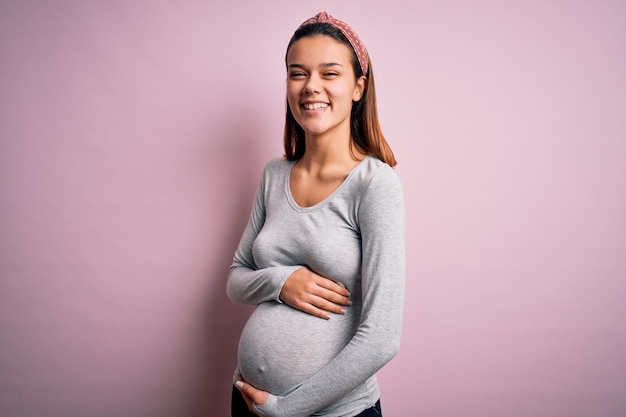 Giovane bella ragazza adolescente incinta in attesa del bambino su sfondo rosa isolato faccia felice sorridente con le braccia incrociate guardando la telecamera Persona positiva