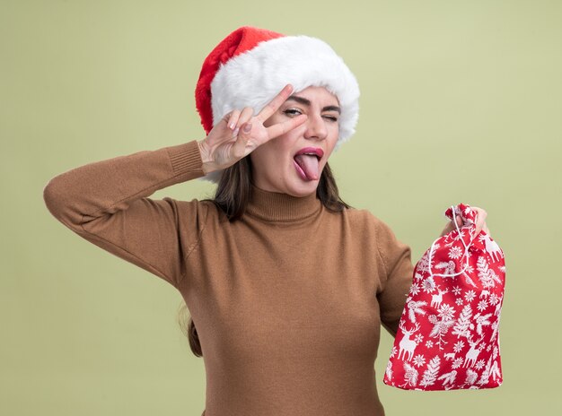 Giovane bella ragazza allegra che porta il cappello di natale che tiene la borsa di natale che mostra la lingua e il gesto di pace isolato sulla parete verde oliva