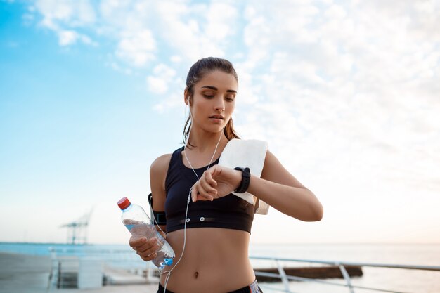 Giovane bella ragazza allegra del brunette che esamina orologio durante l'alba sopra la spiaggia.