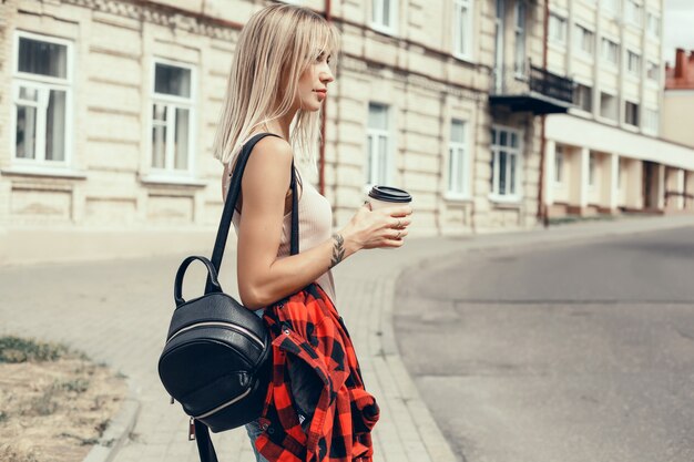 giovane bella ragazza beve il caffè in un bicchiere per strada, ride e sorride