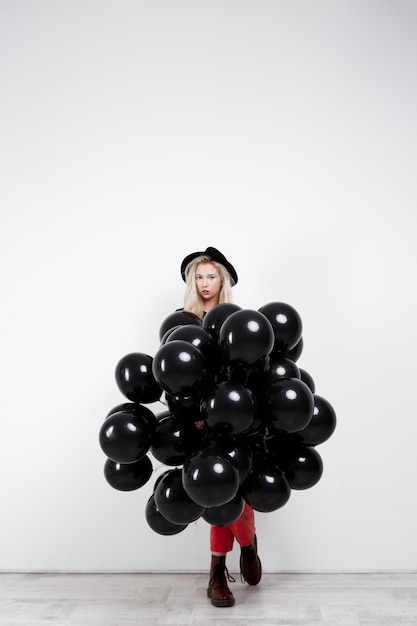 Giovane bella ragazza bionda che tiene baloons neri sopra la parete bianca.