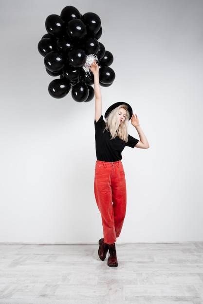 Giovane bella ragazza bionda che tiene baloons neri sopra la parete bianca.