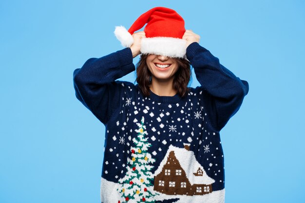 Giovane bella ragazza castana in maglione tricottato e cappello di natale che sorride sopra la parete blu