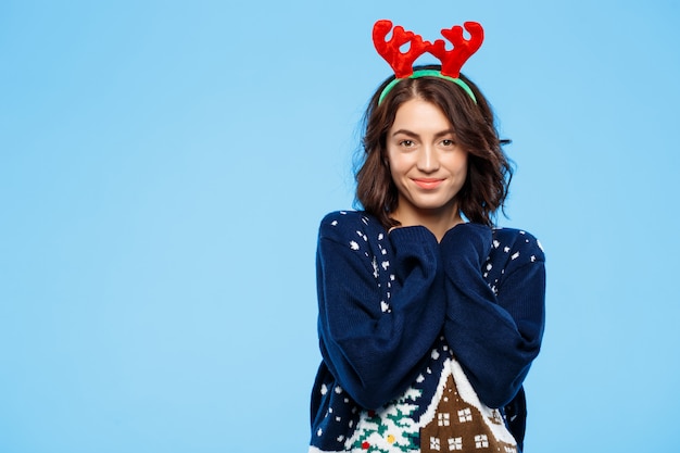 Giovane bella ragazza castana in maglione tricottato e corna di renna di natale che sorridono sopra la parete blu