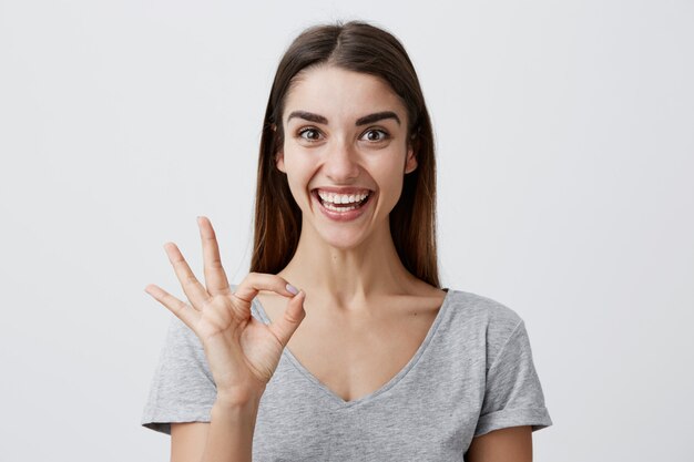 Giovane bella ragazza caucasica allegra con capelli lunghi scuri in camicia grigia casuale che sorride con i denti, facendo segno GIUSTO con le dita, con l'espressione felice ed eccitata del fronte. Copia spazio.