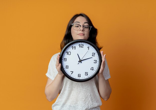 Giovane bella ragazza caucasica con gli occhiali tenendo e guardando l'orologio isolato su sfondo arancione con copia spazio