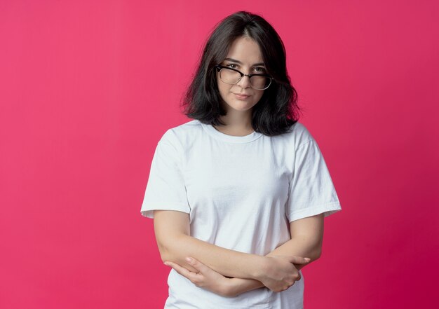 Giovane bella ragazza caucasica con gli occhiali tenendo le mani incrociate sulla pancia guardando la telecamera isolata su sfondo cremisi con copia spazio