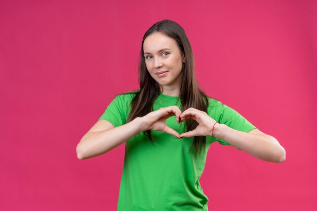 Giovane bella ragazza che indossa la maglietta verde che fa il gesto romantico del cuore sopra il petto sorridente che sta sopra fondo rosa isolato